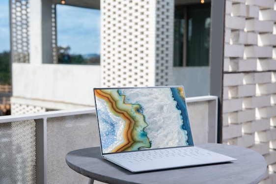 A sleek laptop with a vibrant abstract design on its screen is placed on a round, light grey table. The backdrop includes a modern architectural setting with perforated brick walls and a partially visible glass facade.