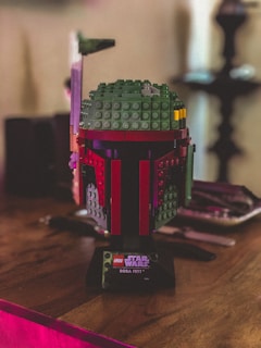 A Lego model of a helmet from Star Wars is placed on a wooden surface. The helmet is constructed with various colored bricks including green, red, and yellow. A plaque at the base reads 'LEGO Star Wars Boba Fett.' The background is blurred, adding focus to the model.