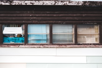 A series of windows with various types of blinds and curtains partially covering them. The window frames appear weathered and have a rustic look, with some visible wear and discoloration. Behind the windows, there are hints of interior objects and perhaps some reflections.