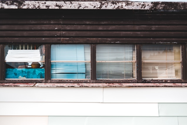 A series of windows with various types of blinds and curtains partially covering them. The window frames appear weathered and have a rustic look, with some visible wear and discoloration. Behind the windows, there are hints of interior objects and perhaps some reflections.