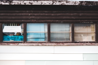 A series of windows with various types of blinds and curtains partially covering them. The window frames appear weathered and have a rustic look, with some visible wear and discoloration. Behind the windows, there are hints of interior objects and perhaps some reflections.
