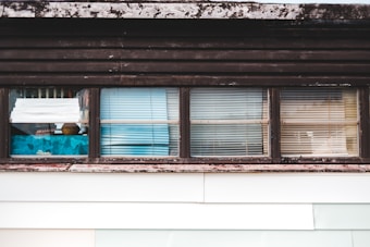 A series of windows with various types of blinds and curtains partially covering them. The window frames appear weathered and have a rustic look, with some visible wear and discoloration. Behind the windows, there are hints of interior objects and perhaps some reflections.