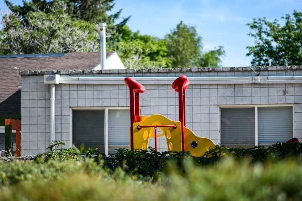 A piece of playground equipment with vibrant red and yellow colors stands in front of a building with a tiled wall and large windows. Lush greenery surrounds the area, and trees are visible in the background under a clear blue sky.