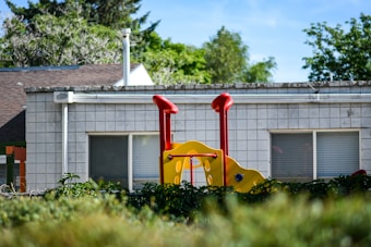 A piece of playground equipment with vibrant red and yellow colors stands in front of a building with a tiled wall and large windows. Lush greenery surrounds the area, and trees are visible in the background under a clear blue sky.