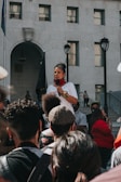 A speaker holds a microphone and addresses a crowd gathered in front of a large, multi-story building. The speaker is wearing a white shirt and colorful bracelets, with a red scarf around the neck. The audience consists of diverse individuals standing and listening attentively. The scene is set outdoors on a sunny day with the building serving as a backdrop.