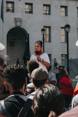 A speaker holds a microphone and addresses a crowd gathered in front of a large, multi-story building. The speaker is wearing a white shirt and colorful bracelets, with a red scarf around the neck. The audience consists of diverse individuals standing and listening attentively. The scene is set outdoors on a sunny day with the building serving as a backdrop.