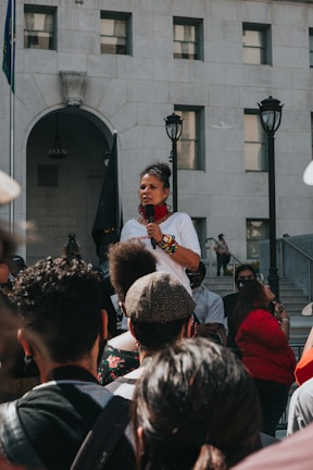 A speaker holds a microphone and addresses a crowd gathered in front of a large, multi-story building. The speaker is wearing a white shirt and colorful bracelets, with a red scarf around the neck. The audience consists of diverse individuals standing and listening attentively. The scene is set outdoors on a sunny day with the building serving as a backdrop.
