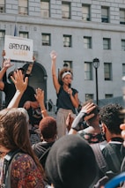 A woman holds a microphone and raises her hand amid a crowd during a protest in front of a building with the word 'JUSTICE' visible. A person nearby holds a sign with the name 'Brendon Glenn' written on it. The crowd is diverse, and many people are raising their hands in solidarity.
