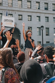 A woman holds a microphone and raises her hand amid a crowd during a protest in front of a building with the word 'JUSTICE' visible. A person nearby holds a sign with the name 'Brendon Glenn' written on it. The crowd is diverse, and many people are raising their hands in solidarity.