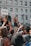 A woman holds a microphone and raises her hand amid a crowd during a protest in front of a building with the word 'JUSTICE' visible. A person nearby holds a sign with the name 'Brendon Glenn' written on it. The crowd is diverse, and many people are raising their hands in solidarity.