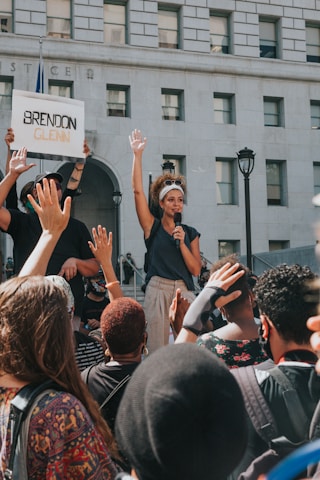 A woman holds a microphone and raises her hand amid a crowd during a protest in front of a building with the word 'JUSTICE' visible. A person nearby holds a sign with the name 'Brendon Glenn' written on it. The crowd is diverse, and many people are raising their hands in solidarity.