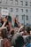 A woman holds a microphone and raises her hand amid a crowd during a protest in front of a building with the word 'JUSTICE' visible. A person nearby holds a sign with the name 'Brendon Glenn' written on it. The crowd is diverse, and many people are raising their hands in solidarity.