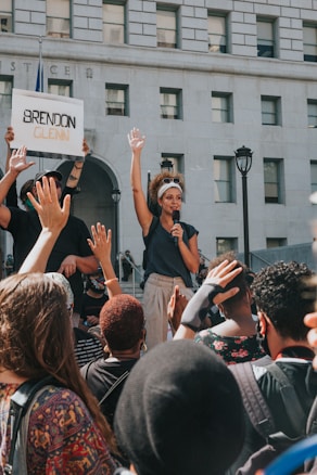 A woman holds a microphone and raises her hand amid a crowd during a protest in front of a building with the word 'JUSTICE' visible. A person nearby holds a sign with the name 'Brendon Glenn' written on it. The crowd is diverse, and many people are raising their hands in solidarity.
