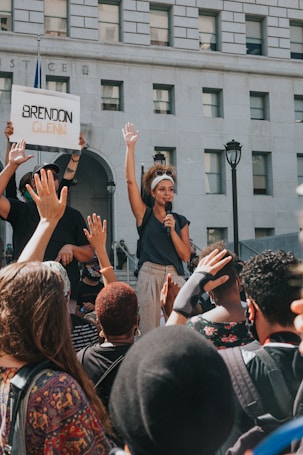 A woman holds a microphone and raises her hand amid a crowd during a protest in front of a building with the word 'JUSTICE' visible. A person nearby holds a sign with the name 'Brendon Glenn' written on it. The crowd is diverse, and many people are raising their hands in solidarity.