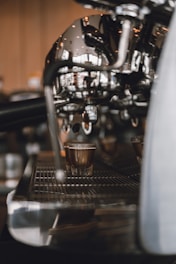 A coffee machine is pouring espresso into a small glass on a metal drip tray. The focus is on the shiny metal parts of the machine, creating a reflective and sleek appearance. The background is softly blurred, highlighting the espresso shot in progress.