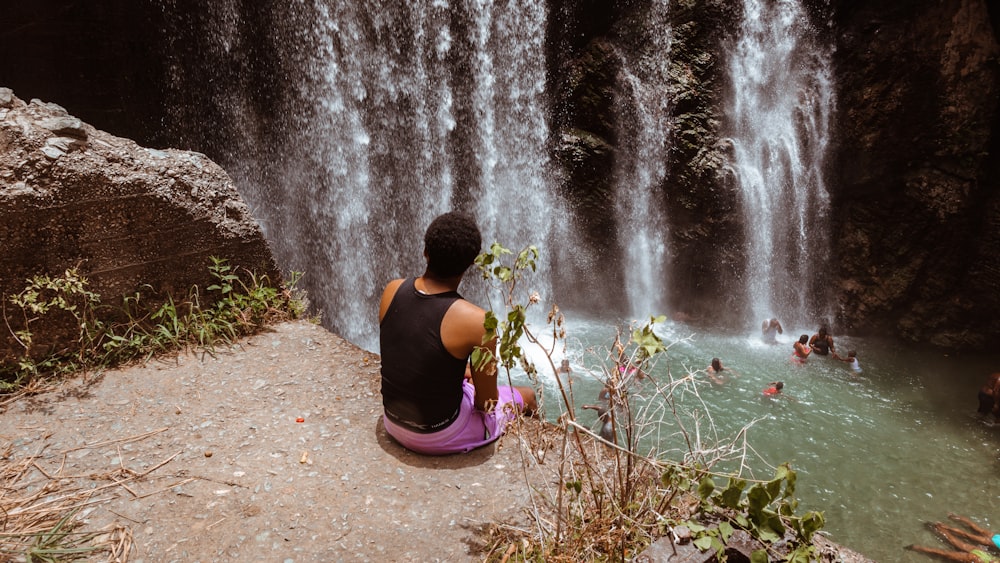 woman in black tank top and purple pants sitting on brown sand near water falls during