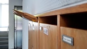 A row of wooden mailboxes, with one open slot holding a large brown envelope protruding outward. The background features a staircase and an elevator door, suggesting a residential or office environment.