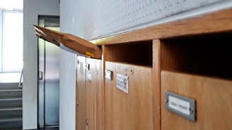 A row of wooden mailboxes, with one open slot holding a large brown envelope protruding outward. The background features a staircase and an elevator door, suggesting a residential or office environment.