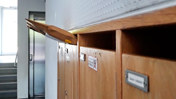A row of wooden mailboxes, with one open slot holding a large brown envelope protruding outward. The background features a staircase and an elevator door, suggesting a residential or office environment.