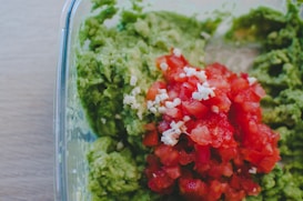A glass dish filled with a mixture of mashed green avocado and chopped red tomatoes topped with bits of chopped white onion or garlic. The colors are vibrant, and the ingredients are fresh.