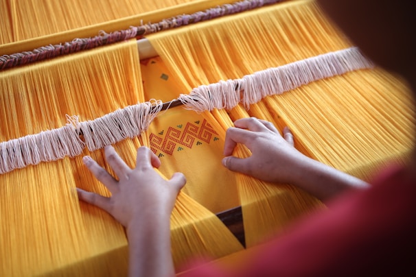 Hands are weaving yellow threads on a loom, creating a textile with an intricate pattern. The textile features a geometric design in orange and red tones. The threads are tightly arranged, and the overall scene suggests careful attention to detail.