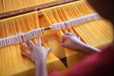 Hands are weaving yellow threads on a loom, creating a textile with an intricate pattern. The textile features a geometric design in orange and red tones. The threads are tightly arranged, and the overall scene suggests careful attention to detail.