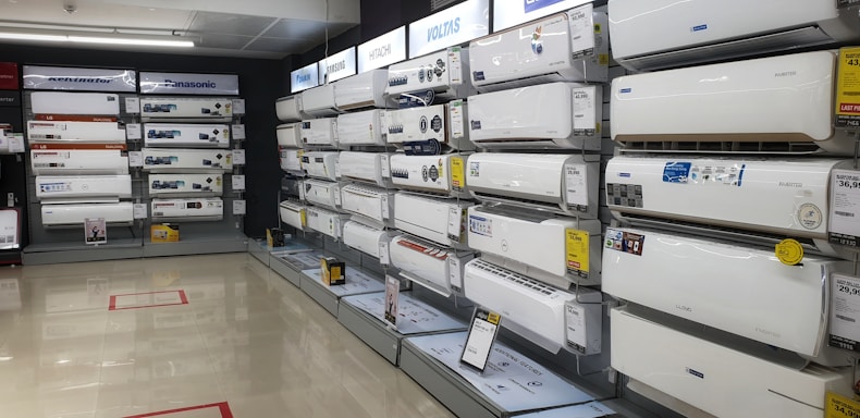 A showroom displaying a variety of air conditioners neatly arranged on two walls. Each unit is placed on shelves with price tags and brand logos visible. The floor is polished and the ceiling has fluorescent lighting.