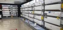 A showroom displaying a variety of air conditioners neatly arranged on two walls. Each unit is placed on shelves with price tags and brand logos visible. The floor is polished and the ceiling has fluorescent lighting.
