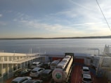 A ferry is carrying several cars, a large tanker truck, and other vehicles on its deck, with a calm body of water and the shoreline visible in the background. The sky is mostly clear with some scattered clouds.