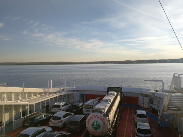 A ferry is carrying several cars, a large tanker truck, and other vehicles on its deck, with a calm body of water and the shoreline visible in the background. The sky is mostly clear with some scattered clouds.