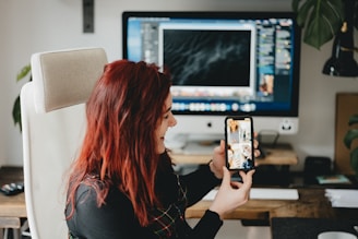A person with red hair is sitting in an office chair, holding a smartphone with a video call or images displayed on the screen. A large computer monitor is on the desk in the background, displaying various windows. The setting is an office or workspace with a wooden desk and a plant nearby.