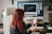 A person with red hair is sitting in an office chair, holding a smartphone with a video call or images displayed on the screen. A large computer monitor is on the desk in the background, displaying various windows. The setting is an office or workspace with a wooden desk and a plant nearby.