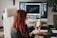 A person with red hair is sitting in an office chair, holding a smartphone with a video call or images displayed on the screen. A large computer monitor is on the desk in the background, displaying various windows. The setting is an office or workspace with a wooden desk and a plant nearby.