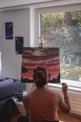 A person with their back facing the viewer is painting a canvas set on an easel. The canvas features a cloudy sky with a full moon and silhouettes of trees and a standing figure, rendered in shades of pink and black. Natural light streams in from a large window, illuminating the scene. Artworks are displayed on the adjacent wall, and there is a sofa and small table in the room.