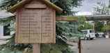 A wooden weather station with a humorous sign describing weather conditions based on the movement or appearance of a hanging rock. The sign is mounted on a wooden post surrounded by greenery and a parked van in the background.