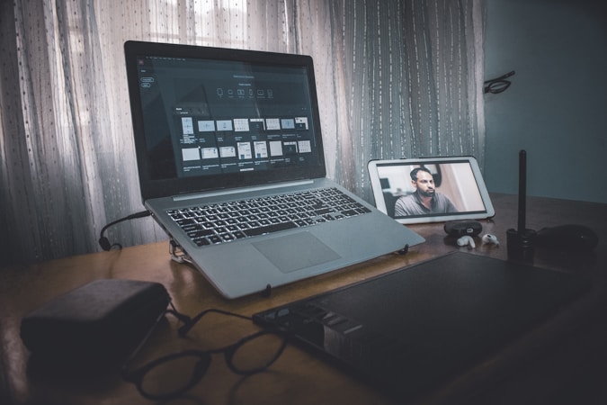 A workspace setup featuring an open laptop displaying design software, with a digital tablet and stylus beside it. A tablet is positioned to the right, showing a video call with a person. Nearby are eyeglasses, earphones, and a small case, all atop a wooden table by a window with sheer curtains letting in soft light.