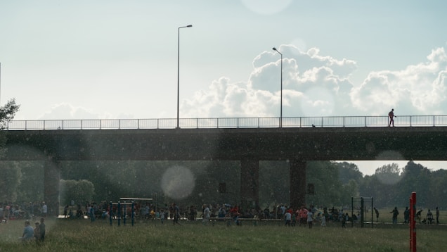 A bridge spans across the top of the image, with a single individual standing on it. Below, a large group of people gathers in what appears to be a park or open space. Trees and grassy fields extend into the background, under a sky filled with fluffy clouds and a light haze. Sunlight filters through the scene, creating lens flare effects and illuminating the gathering.
