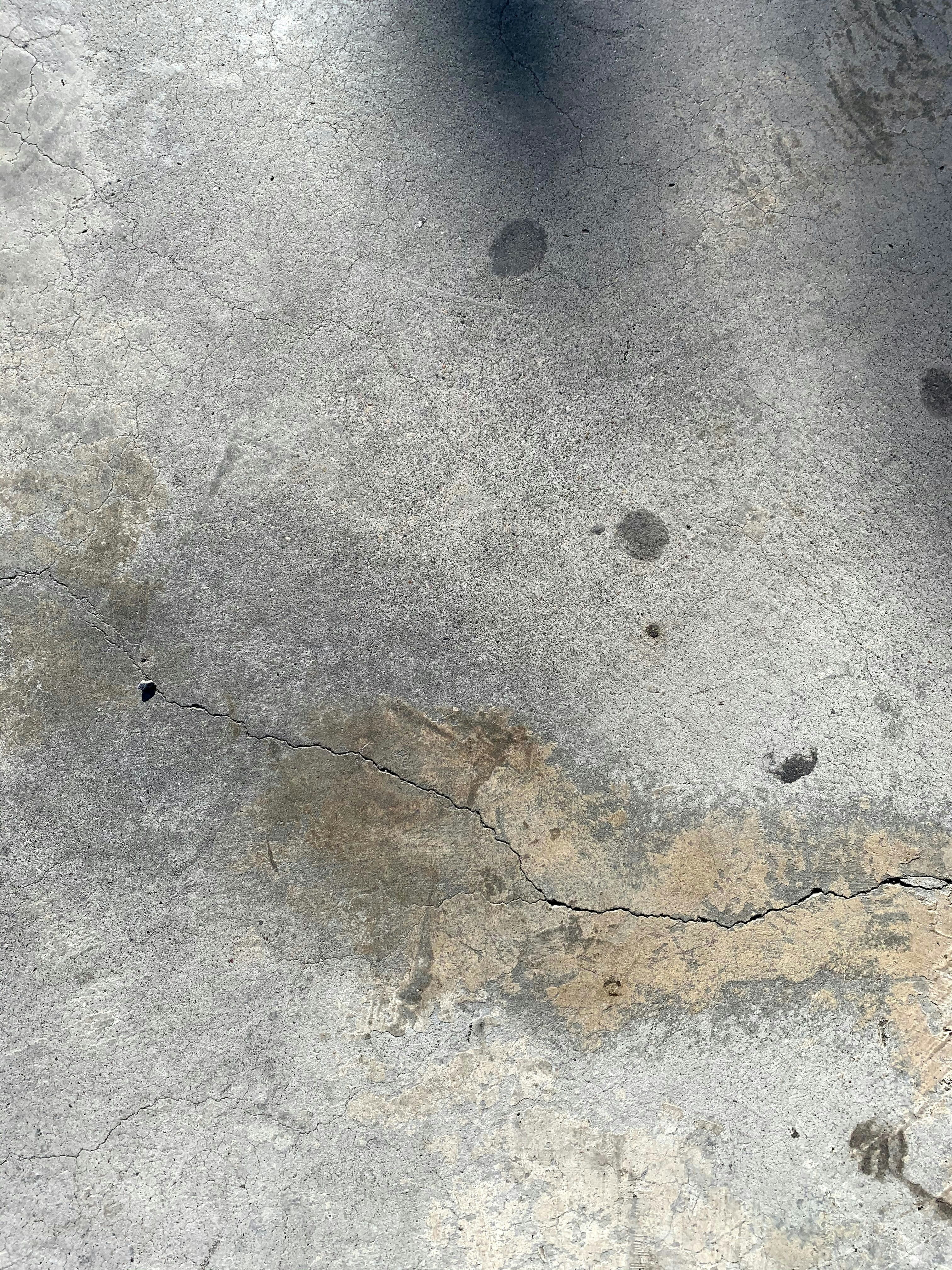 Cracked old concrete driveway with weed growth and deterioration, dim overcast lighting, dark shadows, aged surface