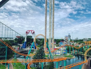 A lively amusement park featuring colorful roller coasters with bright orange and teal tracks. The park includes a loop and various thrill rides situated amidst a lush green landscape and a small body of water. The sky appears partly cloudy, contributing to the vibrant atmosphere.
