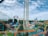 A lively amusement park featuring colorful roller coasters with bright orange and teal tracks. The park includes a loop and various thrill rides situated amidst a lush green landscape and a small body of water. The sky appears partly cloudy, contributing to the vibrant atmosphere.