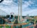 A lively amusement park featuring colorful roller coasters with bright orange and teal tracks. The park includes a loop and various thrill rides situated amidst a lush green landscape and a small body of water. The sky appears partly cloudy, contributing to the vibrant atmosphere.