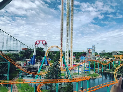 A lively amusement park featuring colorful roller coasters with bright orange and teal tracks. The park includes a loop and various thrill rides situated amidst a lush green landscape and a small body of water. The sky appears partly cloudy, contributing to the vibrant atmosphere.