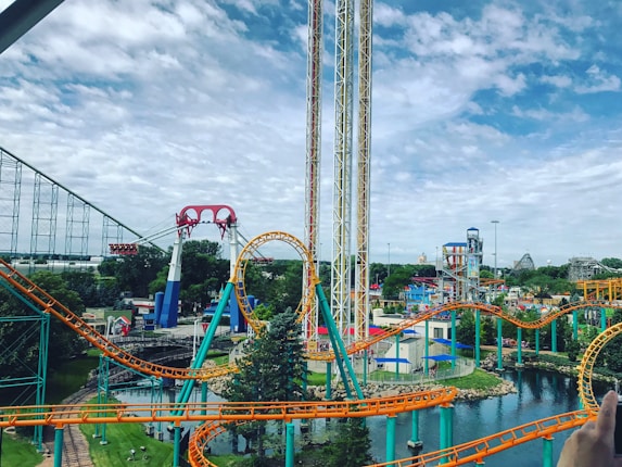 A lively amusement park featuring colorful roller coasters with bright orange and teal tracks. The park includes a loop and various thrill rides situated amidst a lush green landscape and a small body of water. The sky appears partly cloudy, contributing to the vibrant atmosphere.