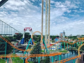A lively amusement park featuring colorful roller coasters with bright orange and teal tracks. The park includes a loop and various thrill rides situated amidst a lush green landscape and a small body of water. The sky appears partly cloudy, contributing to the vibrant atmosphere.