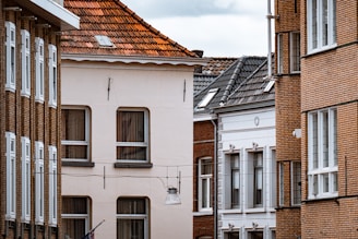 An urban street scene featuring several residential buildings with a mix of architectural styles, including brick and stucco facades. Windows of various designs are prominent across the buildings, with one building having a sloped red-tiled roof. The view is closely cropped, focusing on the textures and details of the walls and windows.
