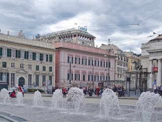 A bustling urban scene with historical architecture featuring buildings with intricate designs and pastel colors. There are several fountains in the foreground, gushing water into the air. A group of people is gathered in the background, possibly tourists or locals. Overcast skies loom above with a hint of cloudiness.