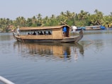 A traditional wooden houseboat with a thatched roof is moving across a calm body of water, surrounded by lush greenery and palm trees. Several people are sitting inside the boat, and there are two individuals at the front and back steering with long wooden poles.