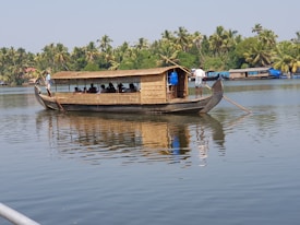 A traditional wooden houseboat with a thatched roof is moving across a calm body of water, surrounded by lush greenery and palm trees. Several people are sitting inside the boat, and there are two individuals at the front and back steering with long wooden poles.