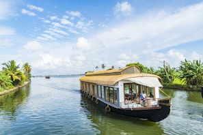 A traditional houseboat sails along a serene river, bordered by lush greenery and palm trees. The bright sky is scattered with fluffy white clouds, casting reflections on the calm water. People are visible on the boat, enjoying the peaceful journey.