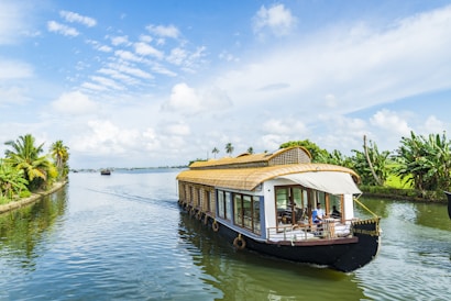 A traditional houseboat sails along a serene river, bordered by lush greenery and palm trees. The bright sky is scattered with fluffy white clouds, casting reflections on the calm water. People are visible on the boat, enjoying the peaceful journey.